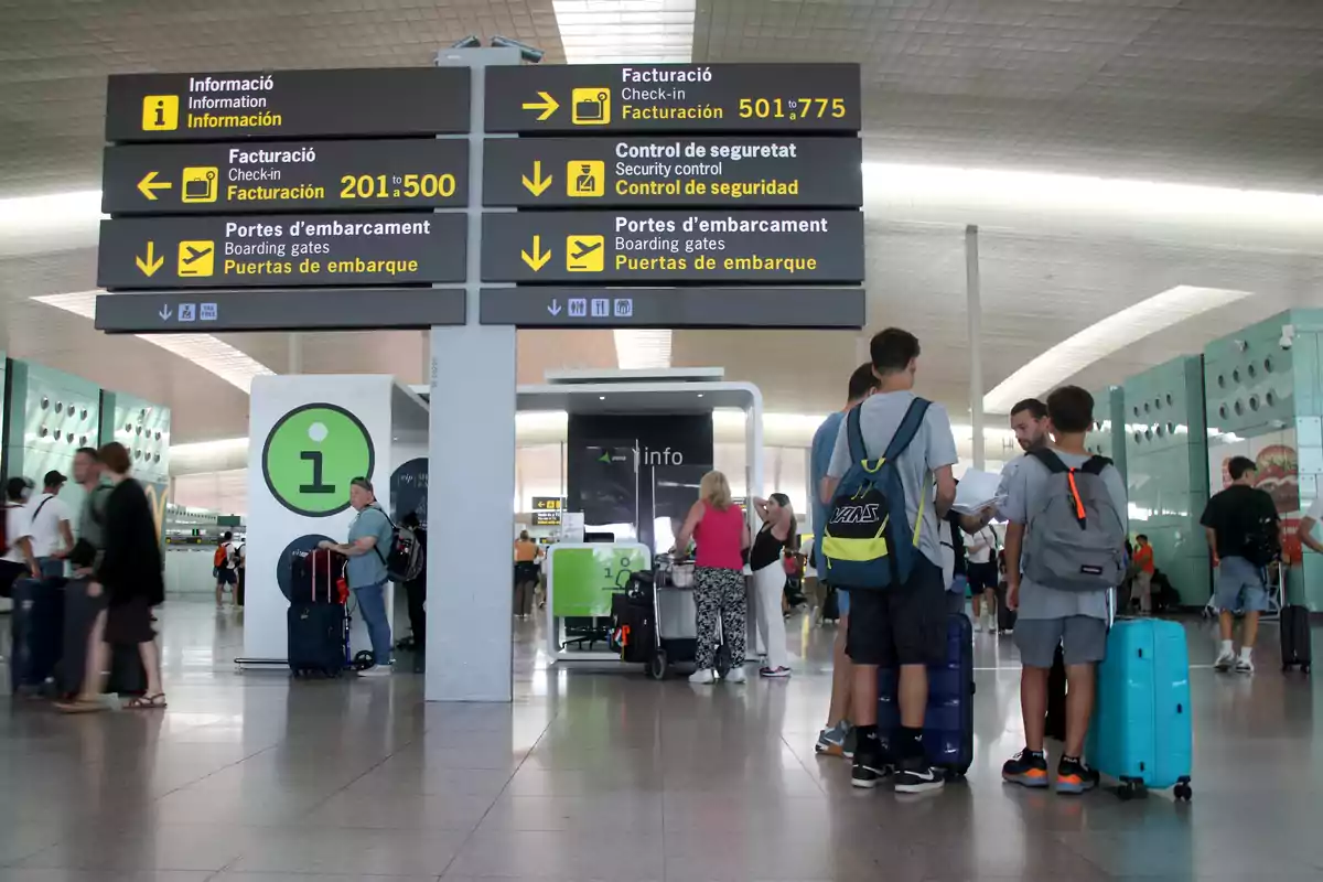 Personas en un aeropuerto observan señales de información, facturación, control de seguridad y puertas de embarque, con maletas y mochilas mientras esperan.