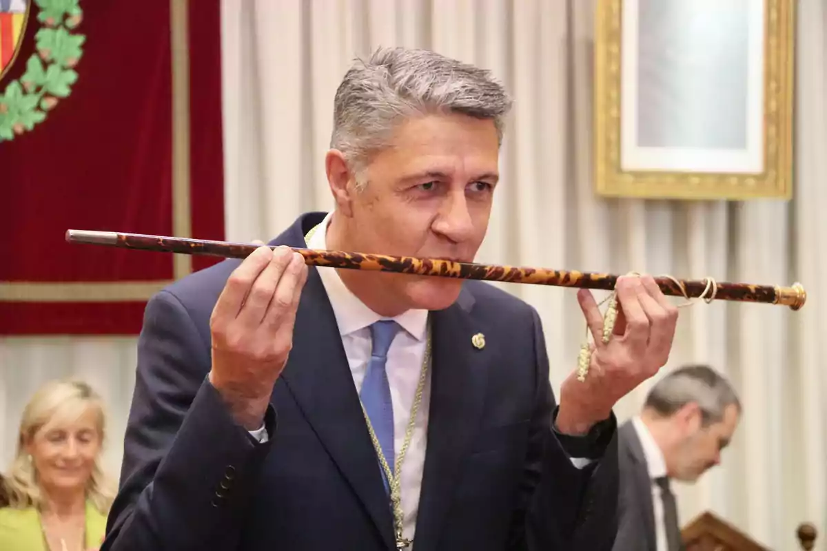 Un hombre en traje besando un bastón ceremonial en un evento formal con personas en el fondo.