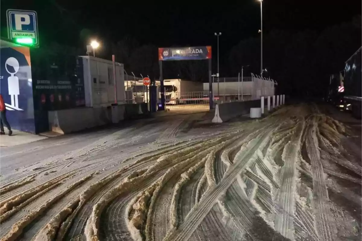 La imagen muestra la entrada de un área de estacionamiento para camiones durante la noche, con un letrero de "Entrada" iluminado y un camino con marcas de neumáticos en la arena.