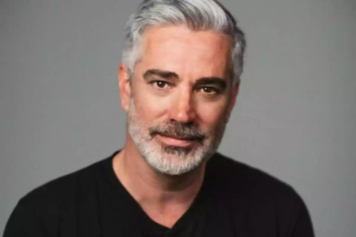 Un hombre de cabello canoso y barba mira a la cámara con una expresión neutral, vistiendo una camiseta negra y con un fondo gris.