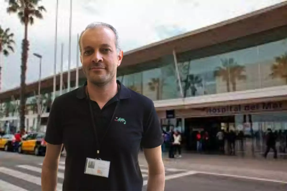 A man in a black T-shirt with an ID card around his neck stands in front of a building with a sign. 