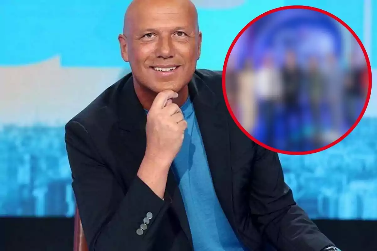 Hombre calvo sonriendo, vestido con una camiseta azul y una chaqueta negra, sentado frente a un fondo azul con una imagen borrosa en un círculo rojo a su derecha.