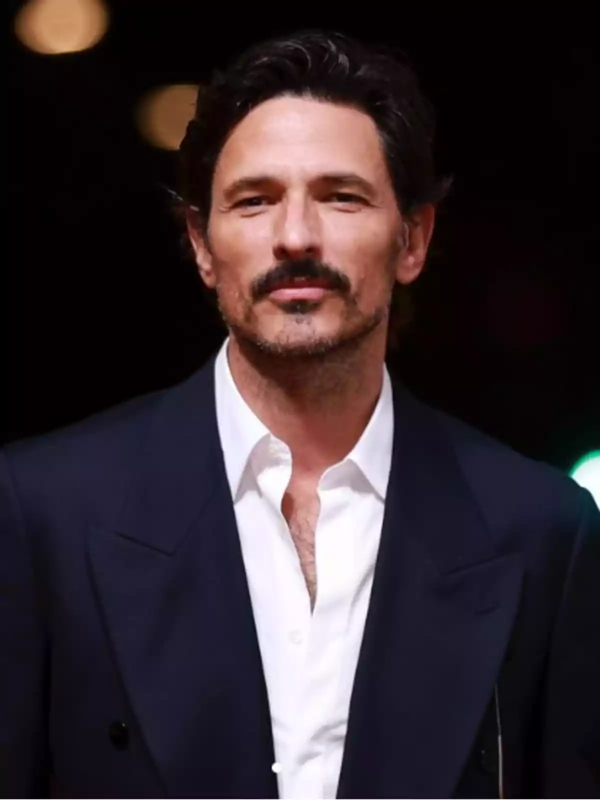 Un hombre con barba y bigote, vestido con un traje oscuro y camisa blanca, posando frente a un fondo oscuro con luces desenfocadas.