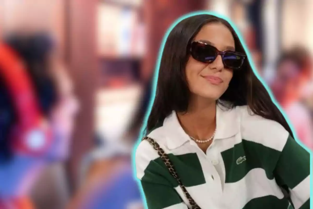 Una mujer con gafas de sol y una camisa de rayas verdes y blancas sonríe mientras está en un entorno interior con un fondo desenfocado.