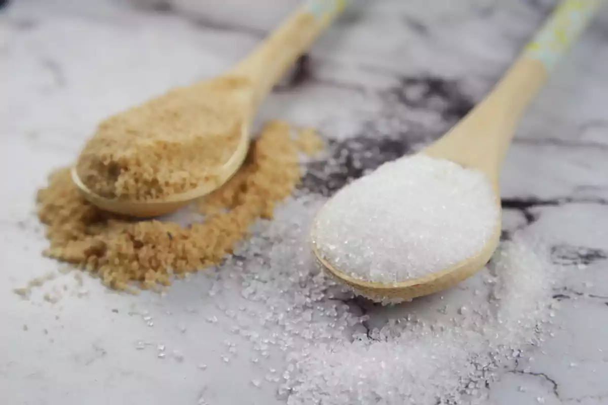 Cucharas de madera con azúcar moreno y azúcar blanco sobre una superficie de mármol.