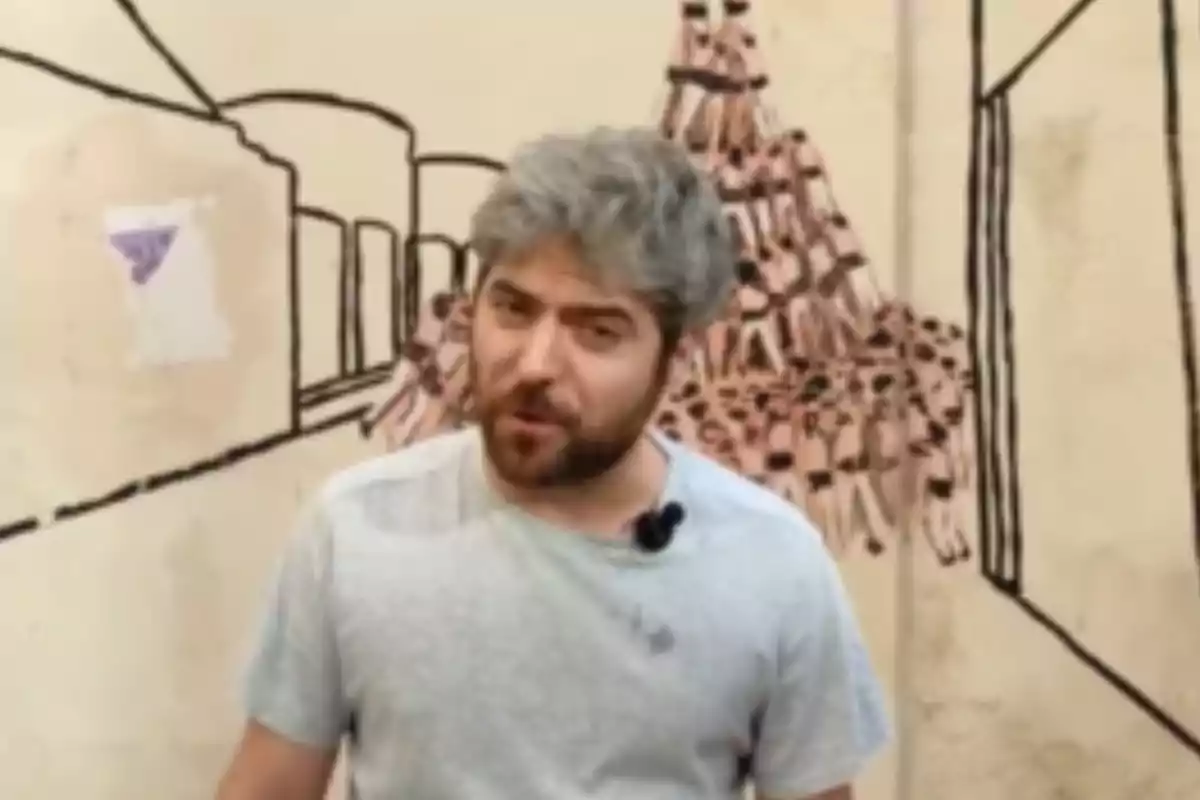 Hombre de cabello canoso y barba, con camiseta gris, hablando frente a un mural con dibujos de edificios y una estructura de botellas.