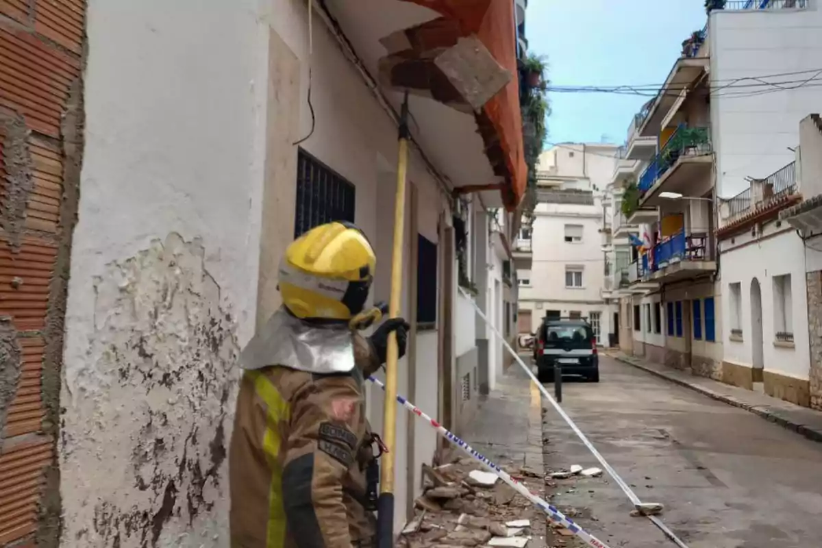 Un bombero con casco amarillo inspecciona un edificio en una calle estrecha mientras sostiene una herramienta larga, con escombros en el suelo y una cinta de seguridad delimitando el área.