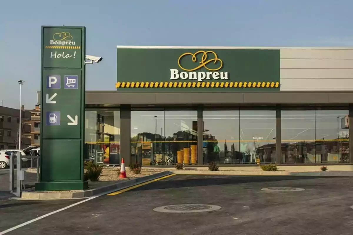 Fachada de un supermercado Bonpreu con un letrero verde que dice "Hola!" y señales de estacionamiento, carrito de compras y gasolinera.