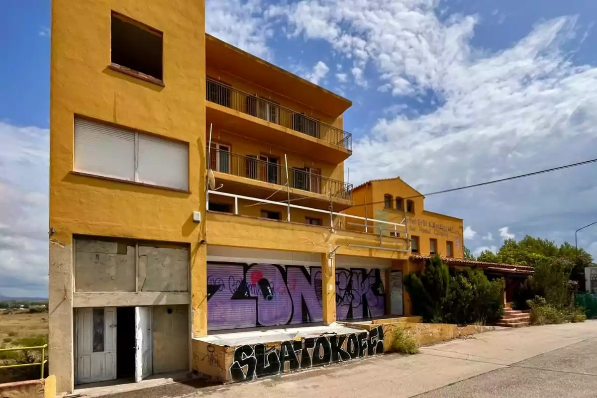 Edificio amarillo abandonado con grafitis en la fachada y cielo parcialmente nublado.