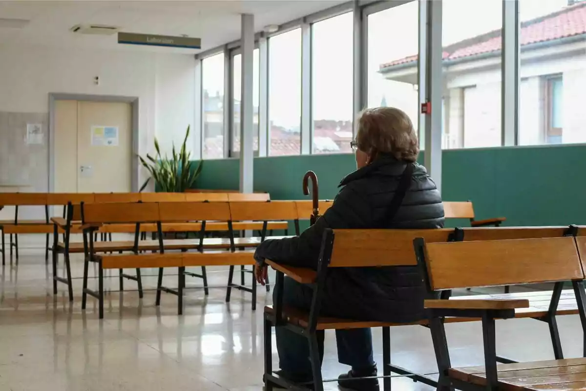 Una persona sentada en una sala de espera vacía con bancos de madera y un bastón en la mano.