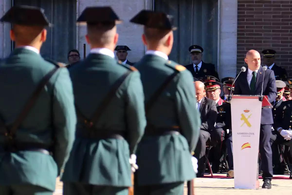 Un grupo de guardias civiles en uniforme verde asisten a un evento oficial al aire libre donde un hombre en traje habla desde un podio.