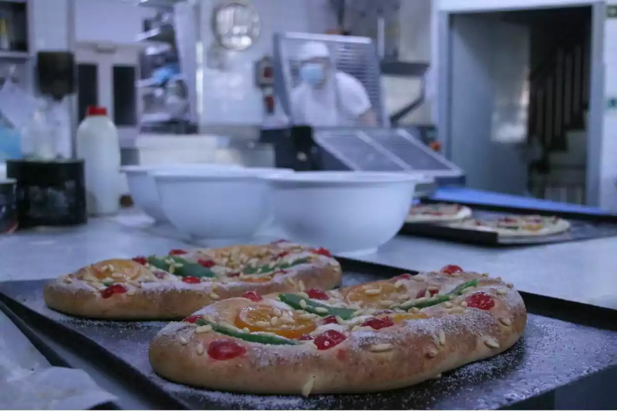 En la imagen se observan dos roscones de reyes recién horneados en una bandeja dentro de una cocina, con un cocinero al fondo usando mascarilla y gorro.