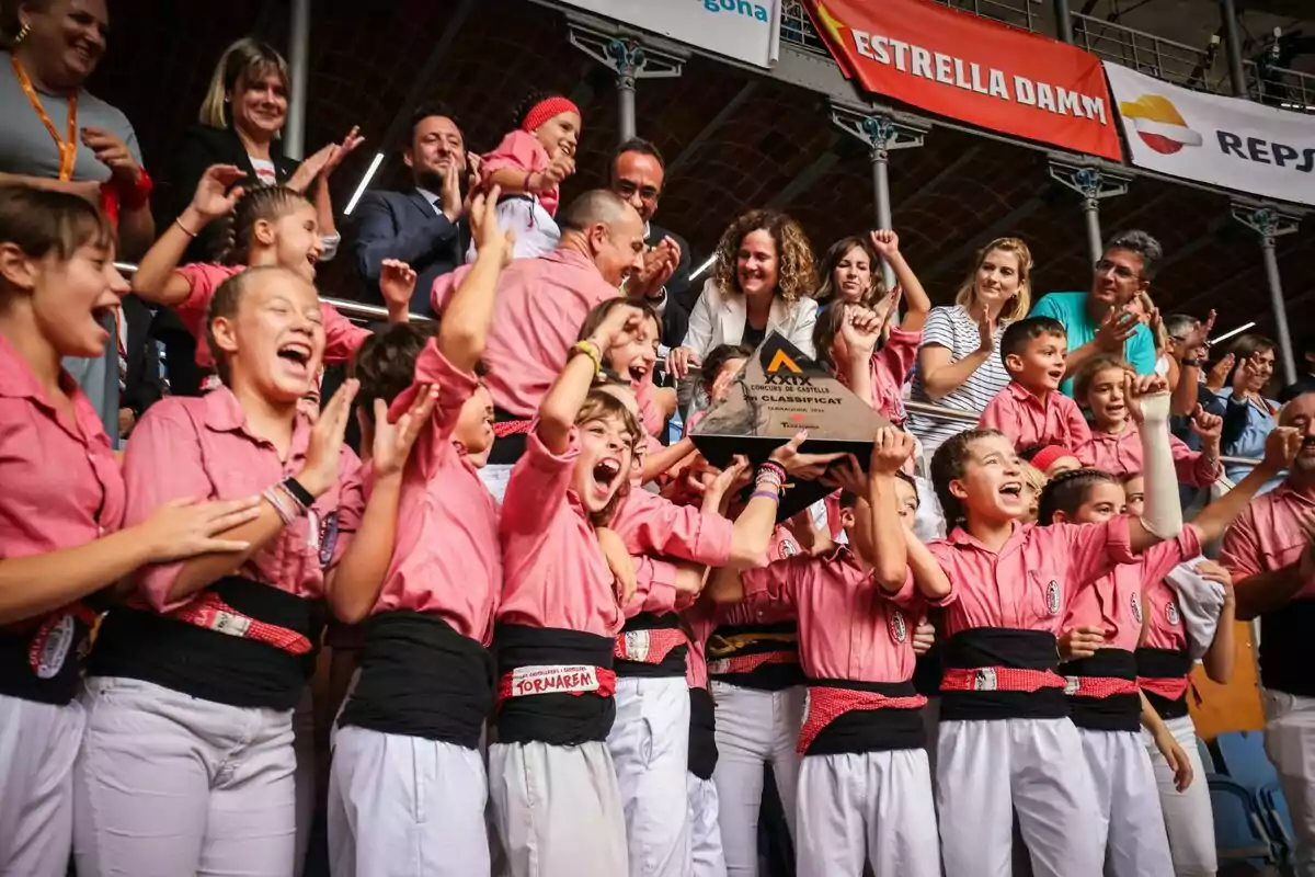 Un grupo de personas celebra con entusiasmo mientras sostienen un trofeo en un evento, todos visten camisas rosadas y pantalones blancos, rodeados de espectadores que aplauden.