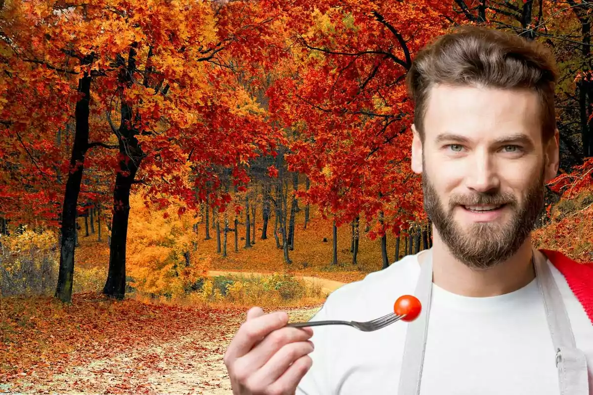 Un hombre con barba sostiene un tenedor con un tomate cherry frente a un paisaje otoñal con árboles de hojas rojas y naranjas.