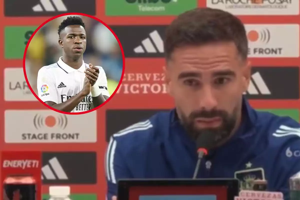 Un hombre con barba y chaqueta deportiva azul está hablando en una conferencia de prensa, mientras que en un círculo rojo se muestra a un jugador de fútbol aplaudiendo.