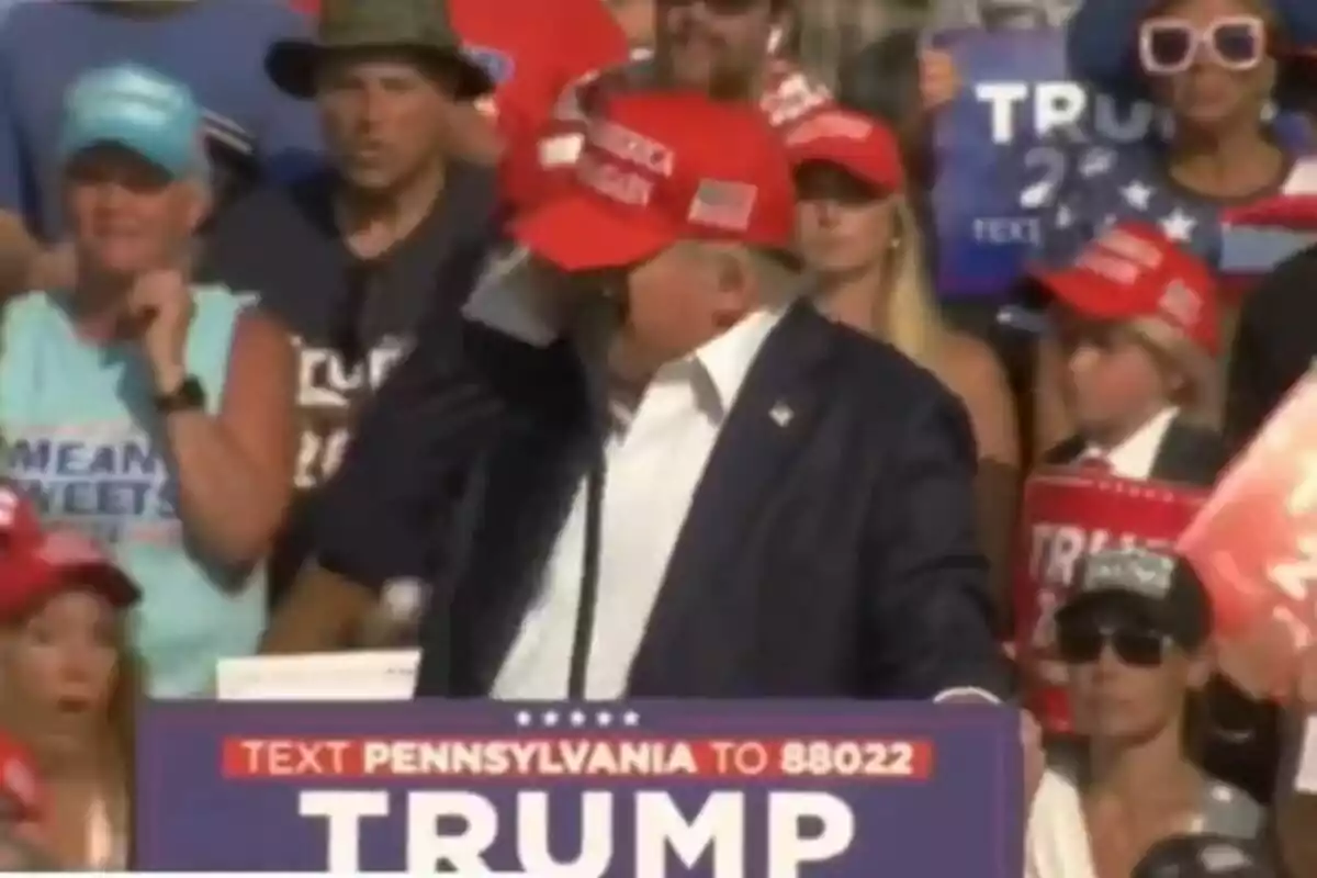 Un hombre con una gorra roja está hablando en un mitin político, rodeado de personas que llevan gorras y camisetas con mensajes de apoyo.