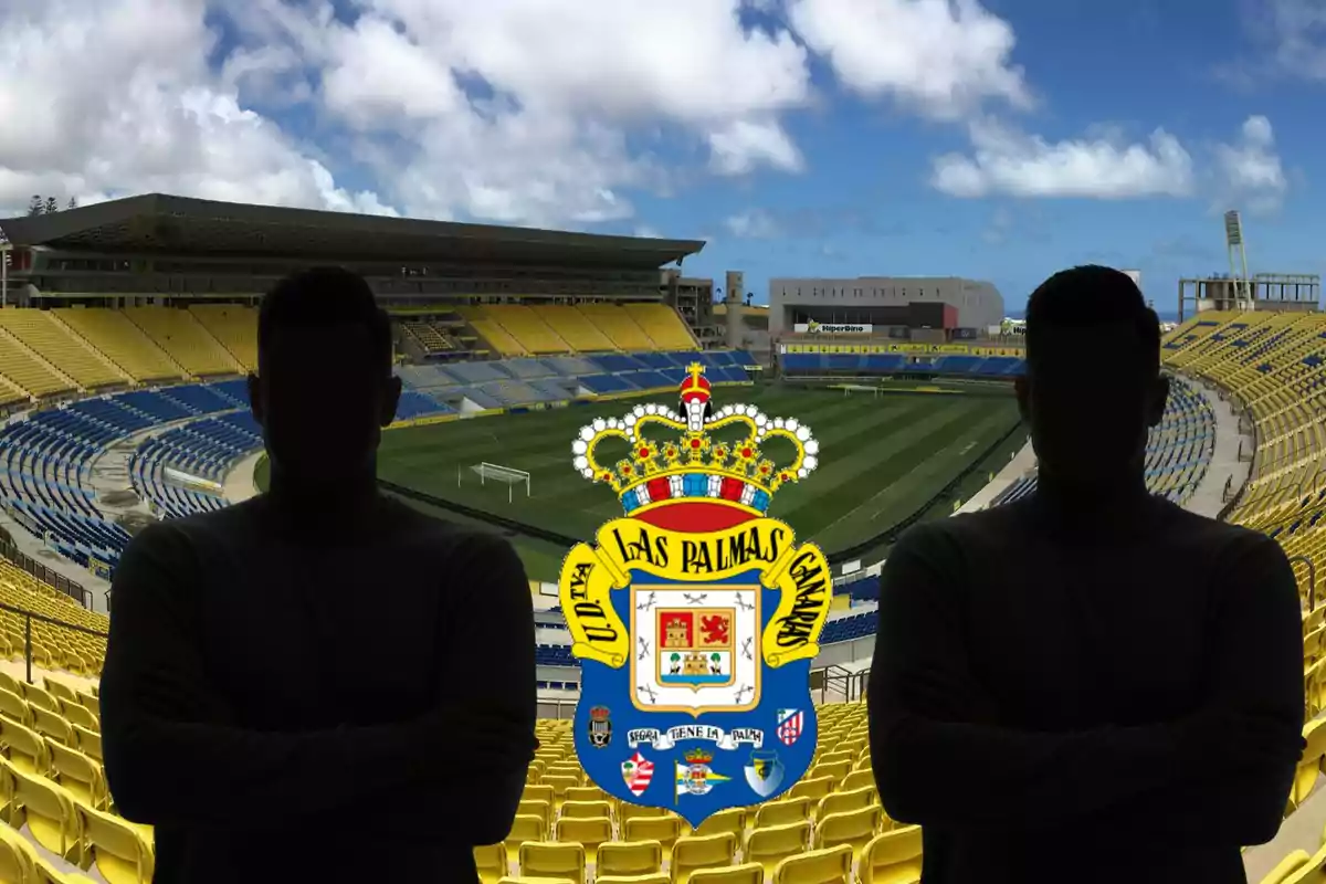 Dos personas con siluetas oscuras posan frente a un estadio de fútbol vacío con asientos amarillos y azules, con el escudo del equipo de fútbol UD Las Palmas en el centro de la imagen.