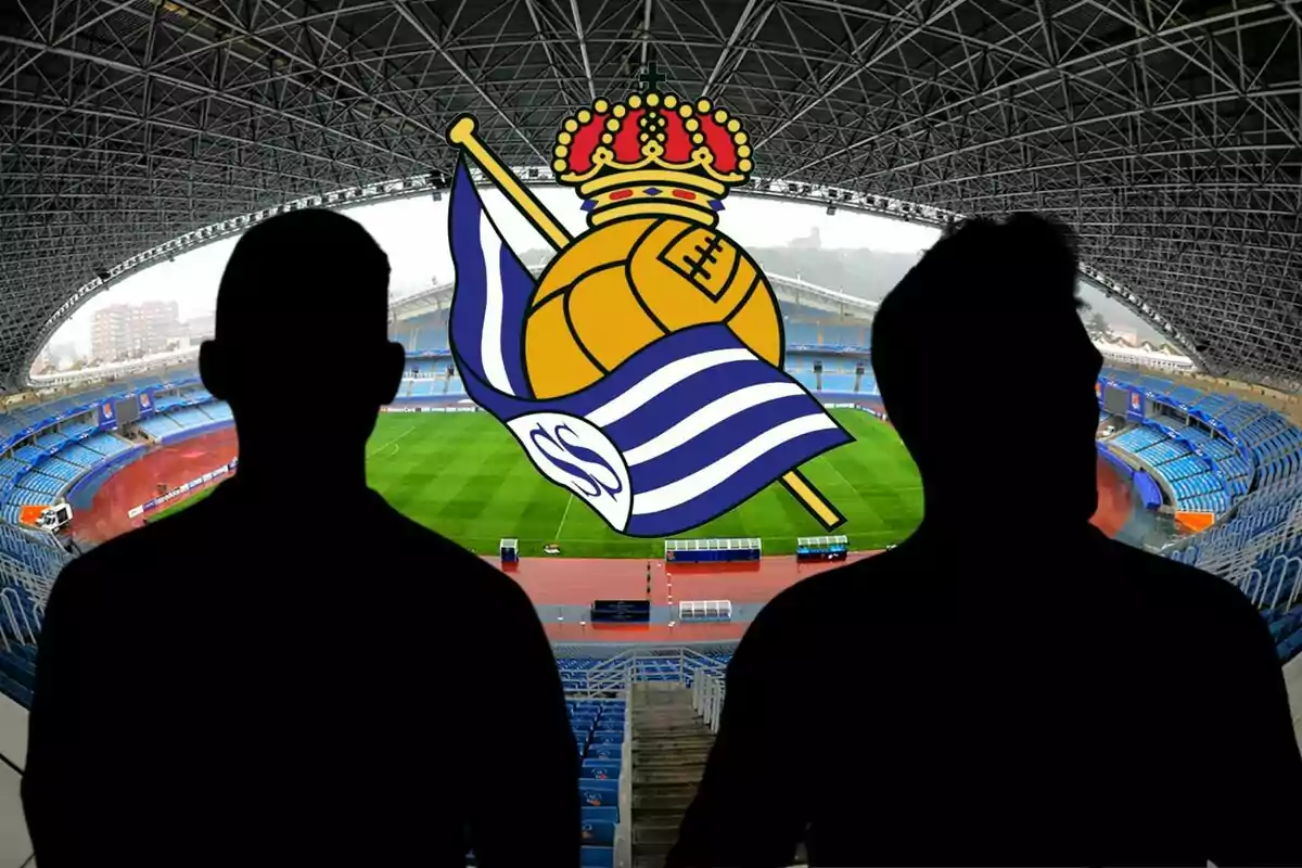 Dos siluetas negras frente a un estadio de fútbol con el escudo de la Real Sociedad en el centro.