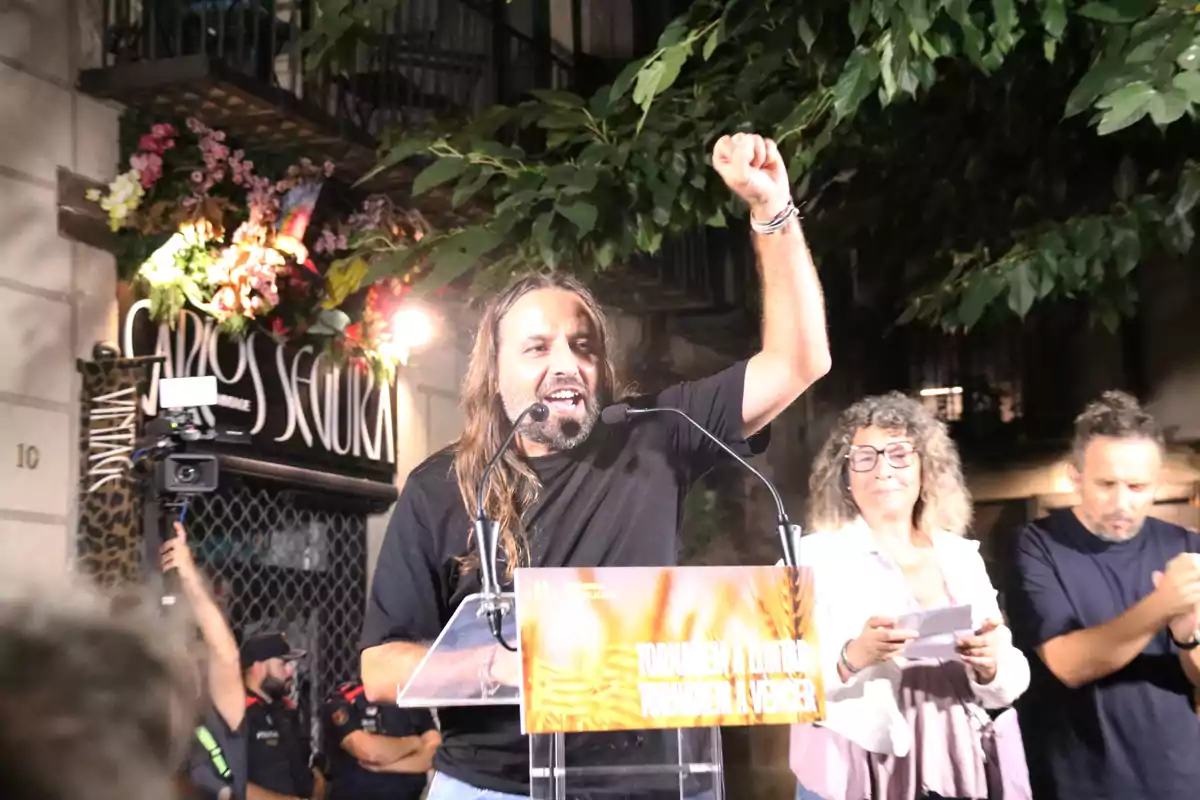 Un hombre con el puño levantado habla en un micrófono durante un evento al aire libre, acompañado por otras dos personas.