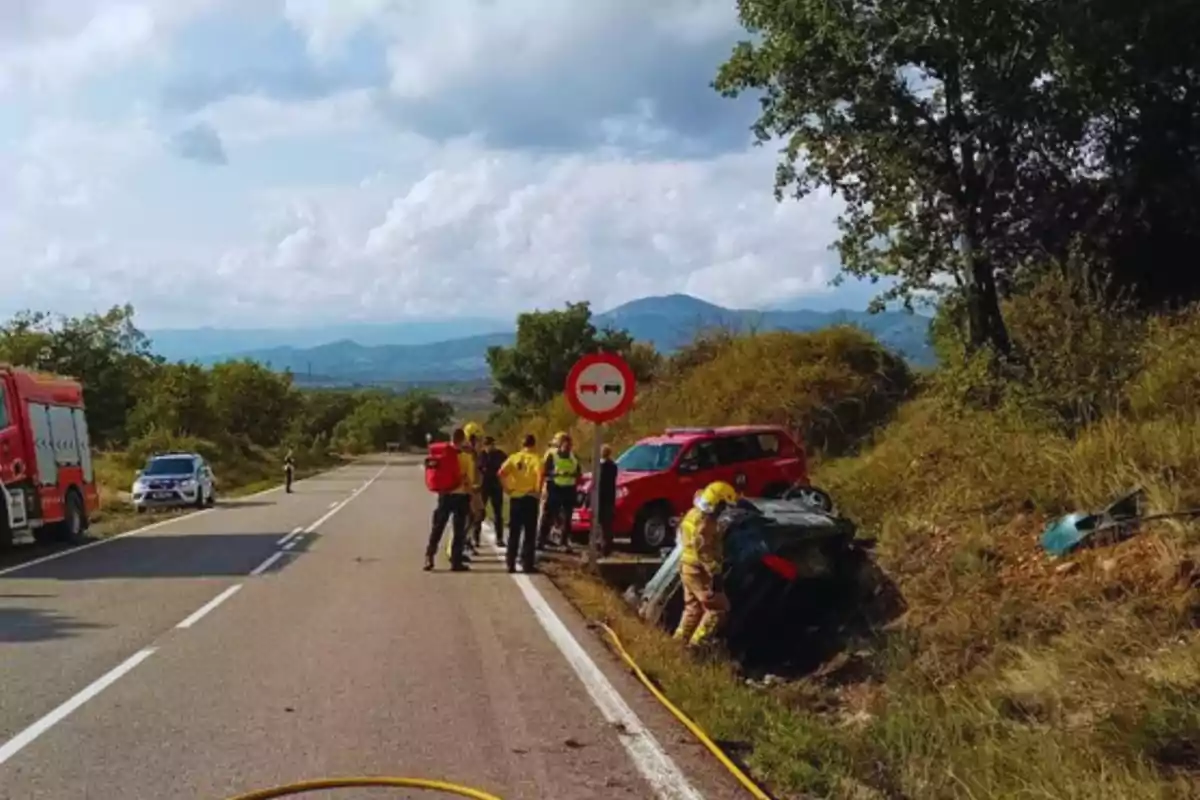 Un grupo de bomberos y rescatistas atienden un accidente de tráfico en una carretera rural, con un coche volcado en la cuneta y vehículos de emergencia en la escena.