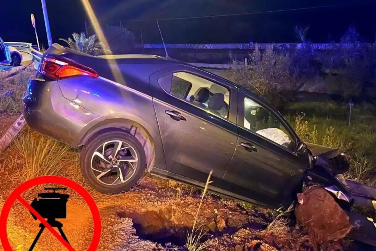 Un coche gris accidentado en una zanja al lado de la carretera durante la noche con luces de emergencia iluminando la escena y un símbolo de prohibición sobre un radar de velocidad en la esquina inferior izquierda.