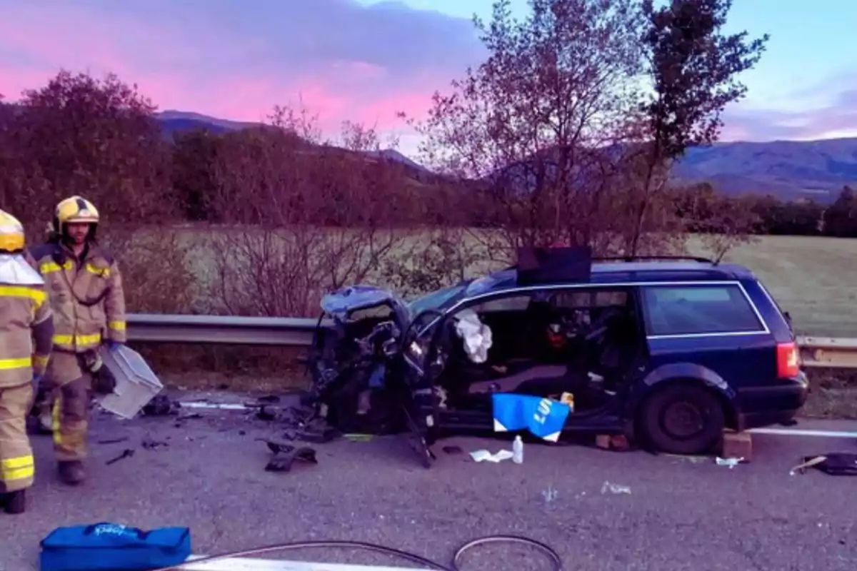 Un coche azul con la parte delantera dañada tras un accidente en una carretera rural al atardecer con dos bomberos en la escena.