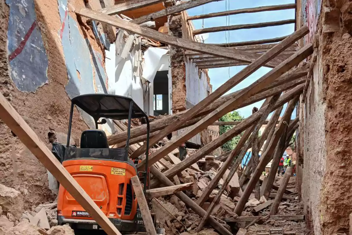 Una excavadora naranja se encuentra en un edificio en ruinas con escombros y vigas de madera caídas.