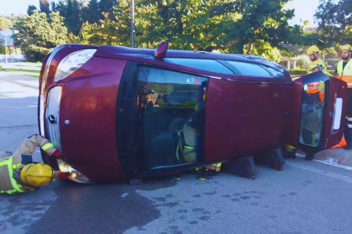 Un coche volcado de lado en la carretera con bomberos trabajando alrededor.