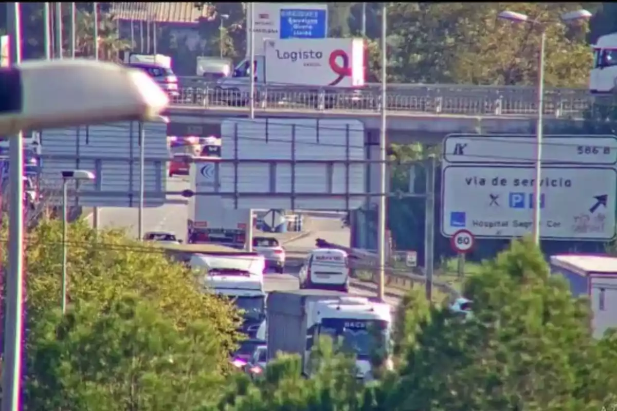 Una carretera con tráfico denso y varios vehículos circulando en ambas direcciones, con un puente elevado y señales de tráfico visibles.