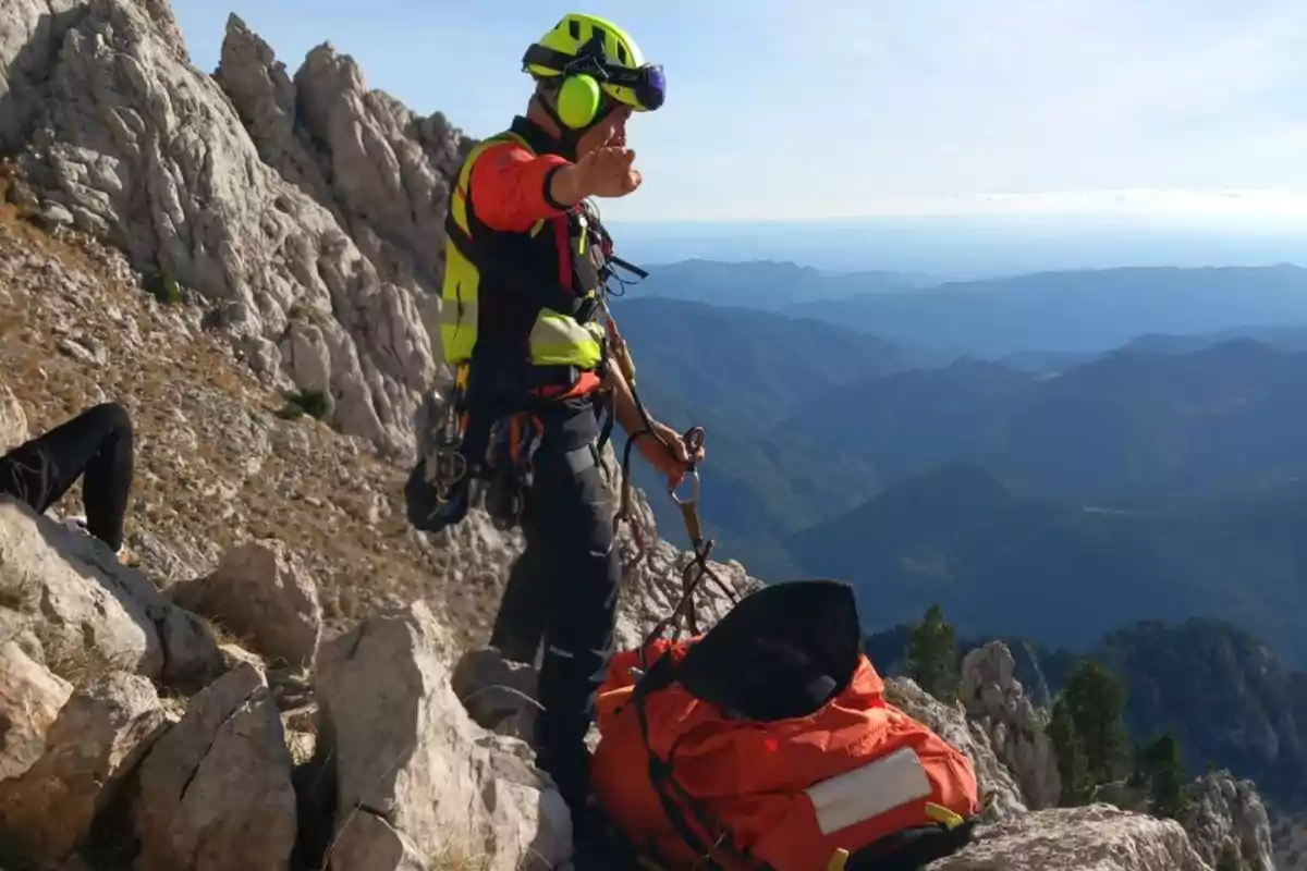 Un rescatista equipado con casco y arnés se encuentra en una montaña rocosa junto a una camilla naranja, con un paisaje montañoso al fondo.