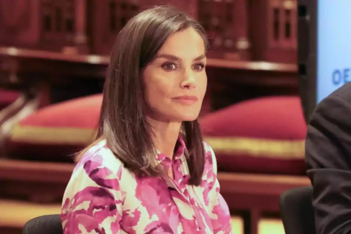 Mujer con cabello castaño y lacio, usando una blusa con estampado floral en tonos rosados, sentada en un entorno interior con decoración de madera y cojines rojos.