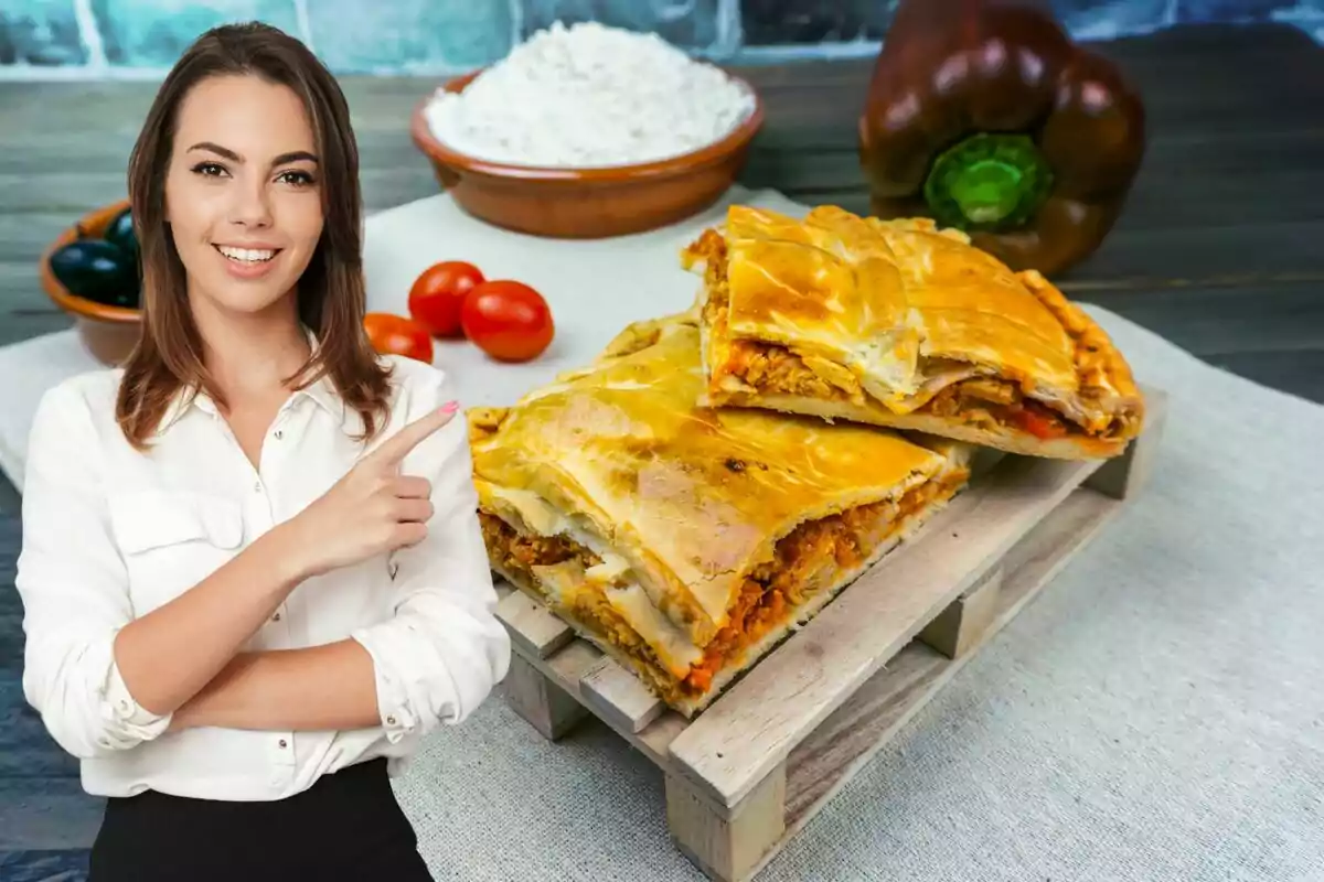 Una mujer sonriente con camisa blanca señala hacia una empanada gallega sobre una mesa, con ingredientes como tomates cherry, pimientos y harina en el fondo.