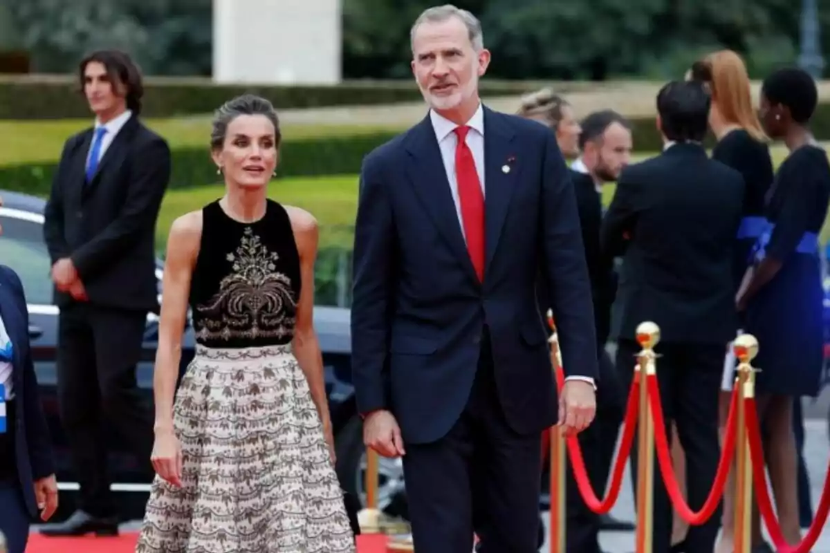 Una pareja elegantemente vestida camina por una alfombra roja, rodeada de personas y cuerdas de terciopelo rojo.