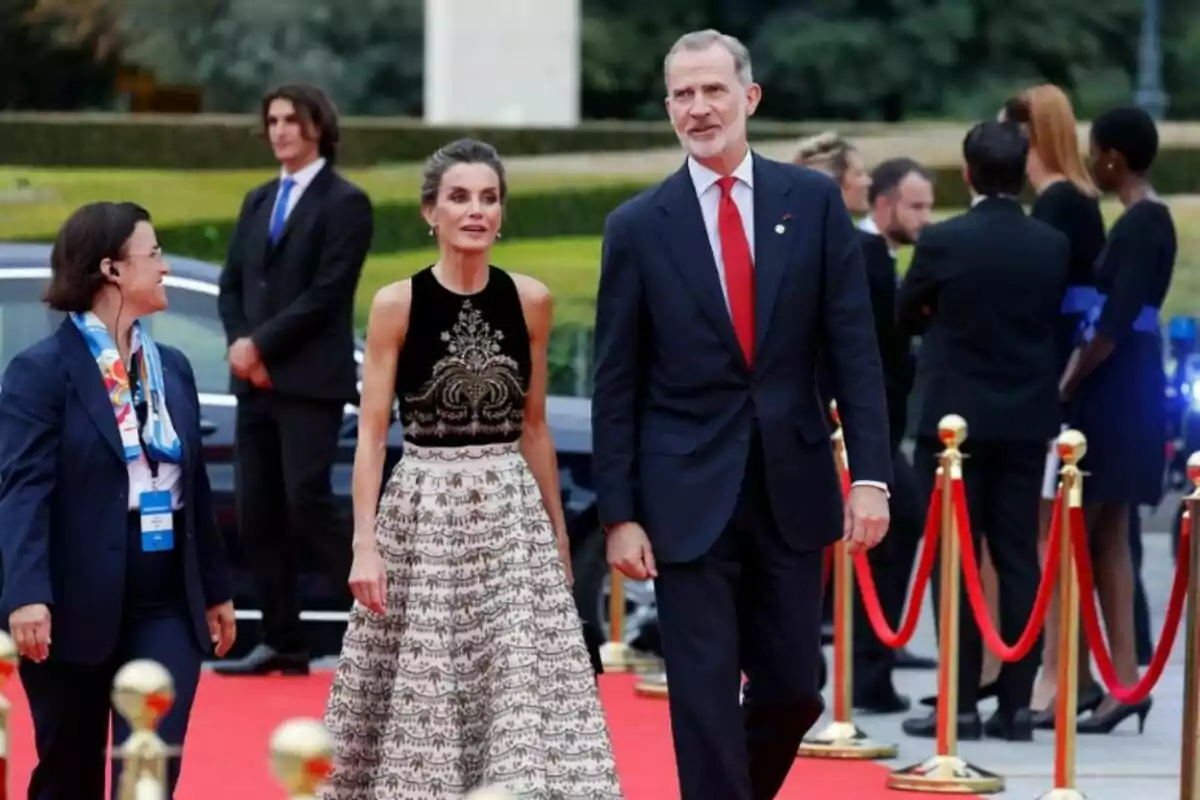 Una pareja elegantemente vestida camina por una alfombra roja, rodeada de otras personas y cuerdas de terciopelo rojo.