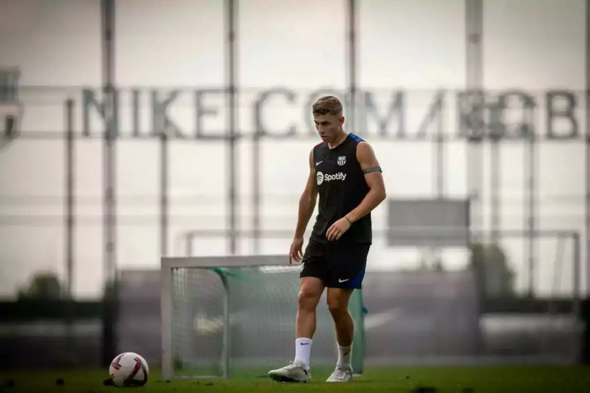 Un jugador de fútbol con uniforme de entrenamiento negro y una pelota en el campo de juego, con una portería y un cartel de Nike en el fondo.