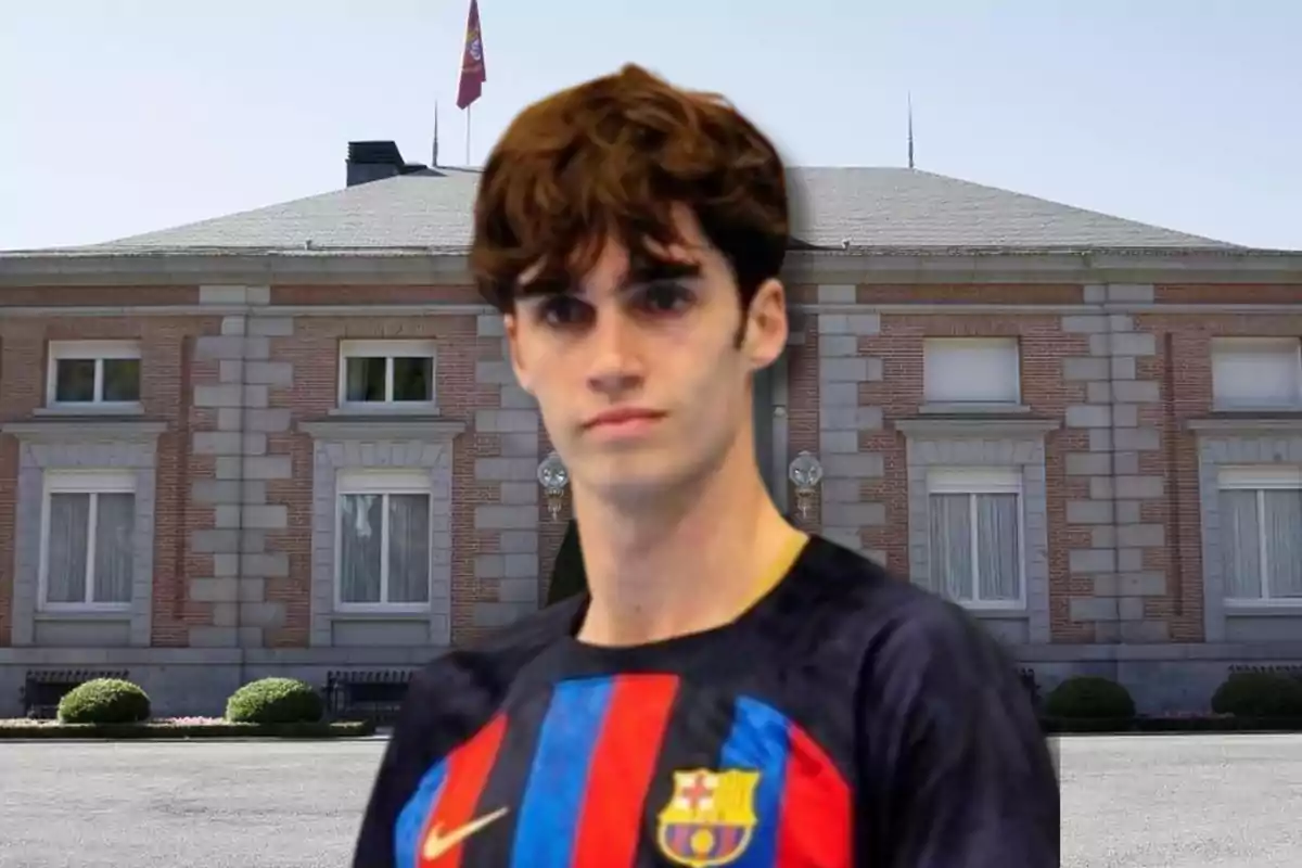 Un joven con una camiseta del FC Barcelona está de pie frente a un edificio de ladrillo con ventanas grandes y una bandera ondeando en el techo.