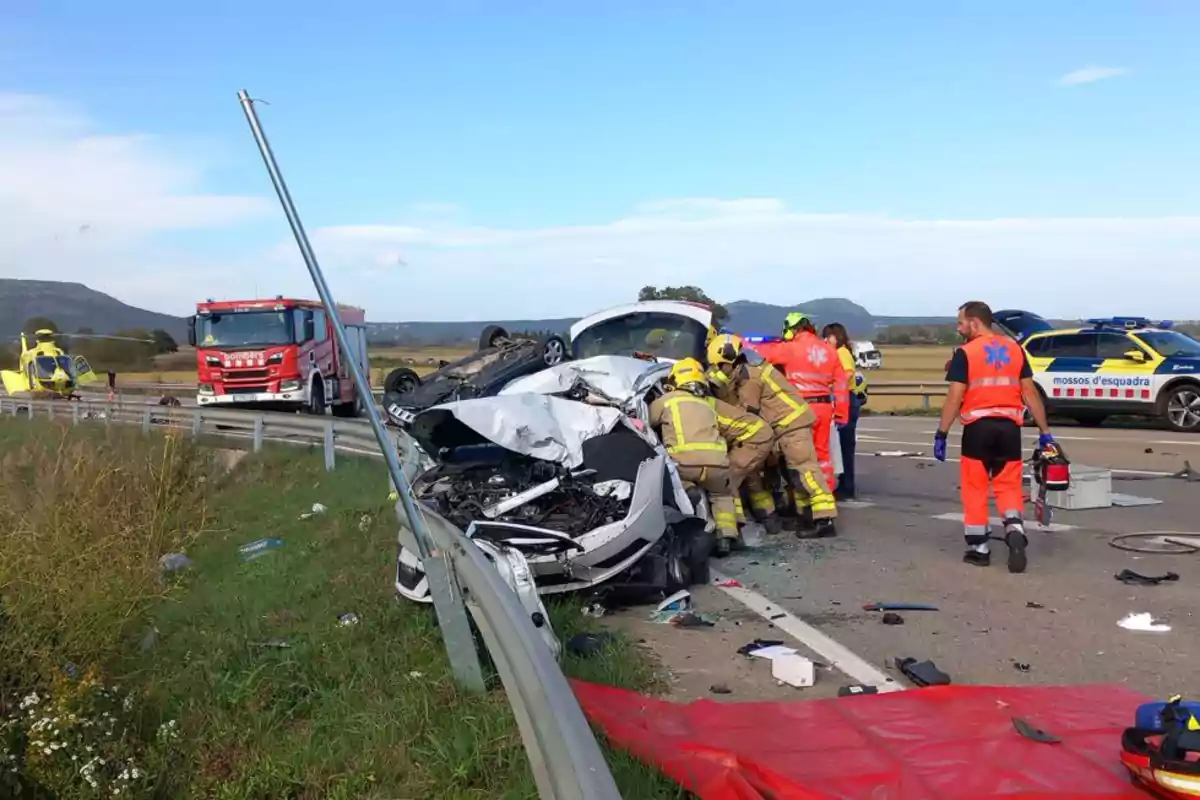 Accidente de tráfico en una carretera con un coche volcado y otro gravemente dañado; bomberos y personal de emergencia trabajando en el lugar, con un camión de bomberos, un helicóptero y un coche de policía presentes.