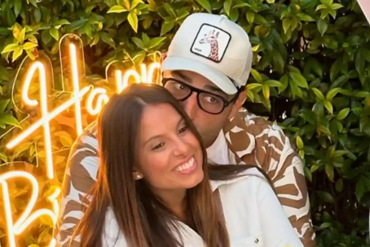 Una pareja sonriente se abraza frente a un letrero de neón que dice "Happy Birthday", rodeados de un fondo de hojas verdes.