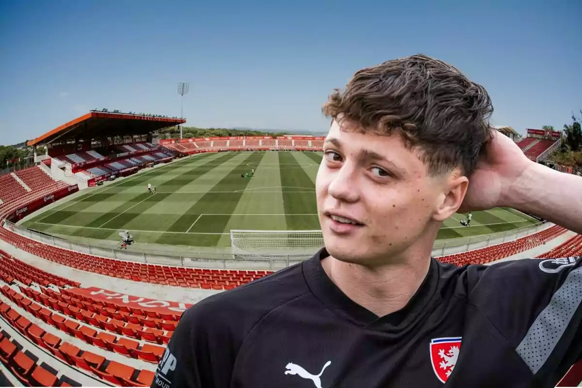 Un jugador de fútbol con una camiseta negra posa frente a un estadio vacío con gradas rojas y un campo de césped bien cuidado.