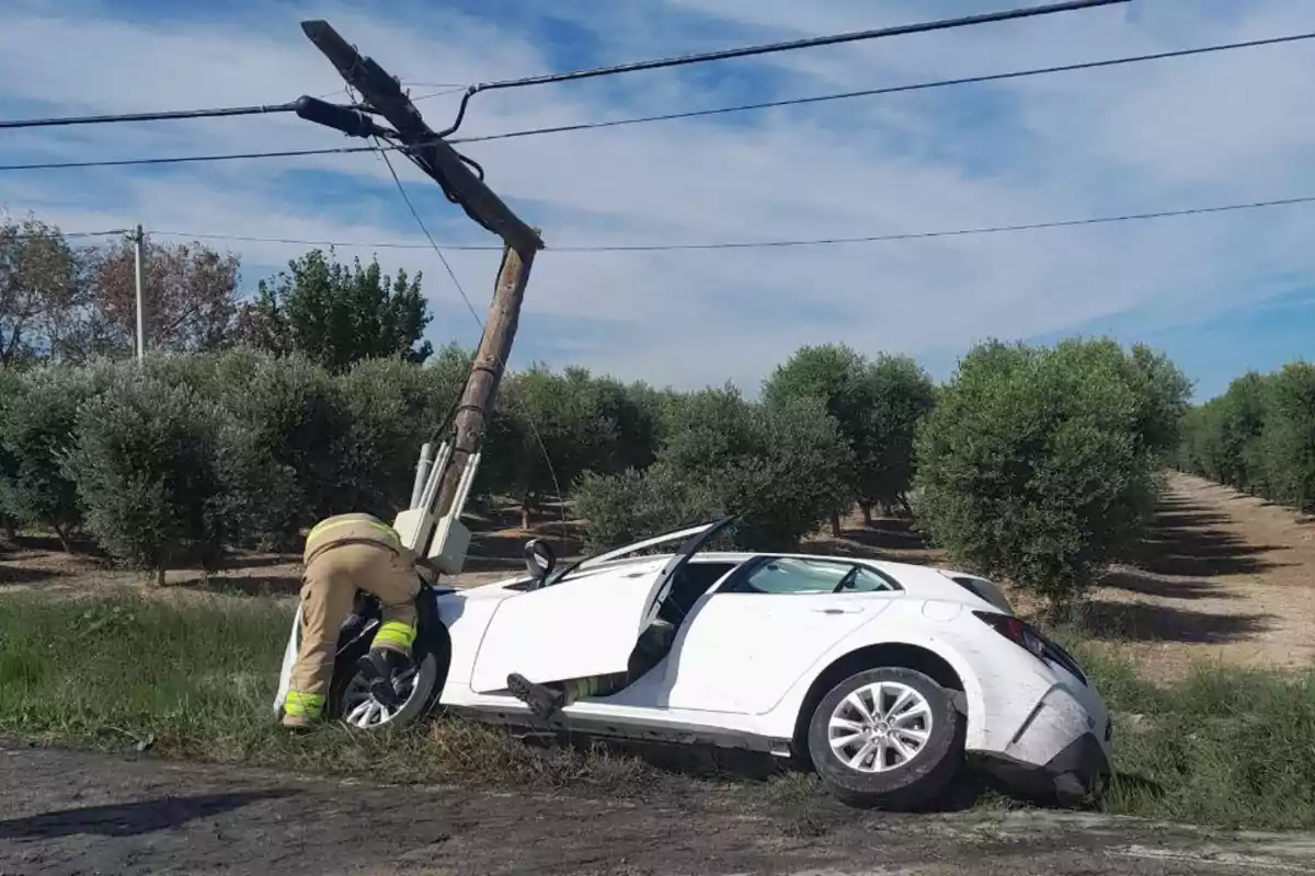 Un coche blanco choca contra un poste de electricidad en una carretera rural mientras un bombero inspecciona el daño.