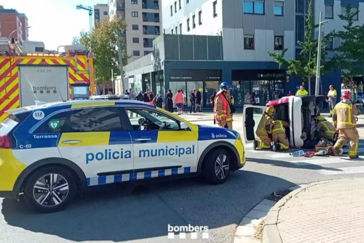 Un coche de la policía municipal y un camión de bomberos en la escena de un accidente de tráfico con un vehículo volcado y bomberos asistiendo.