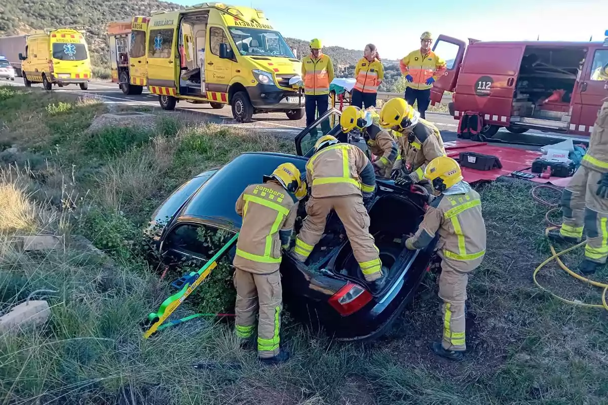 Bomberos y personal de emergencia trabajando en el rescate de un coche accidentado en una zanja con ambulancias y vehículos de emergencia en el fondo.