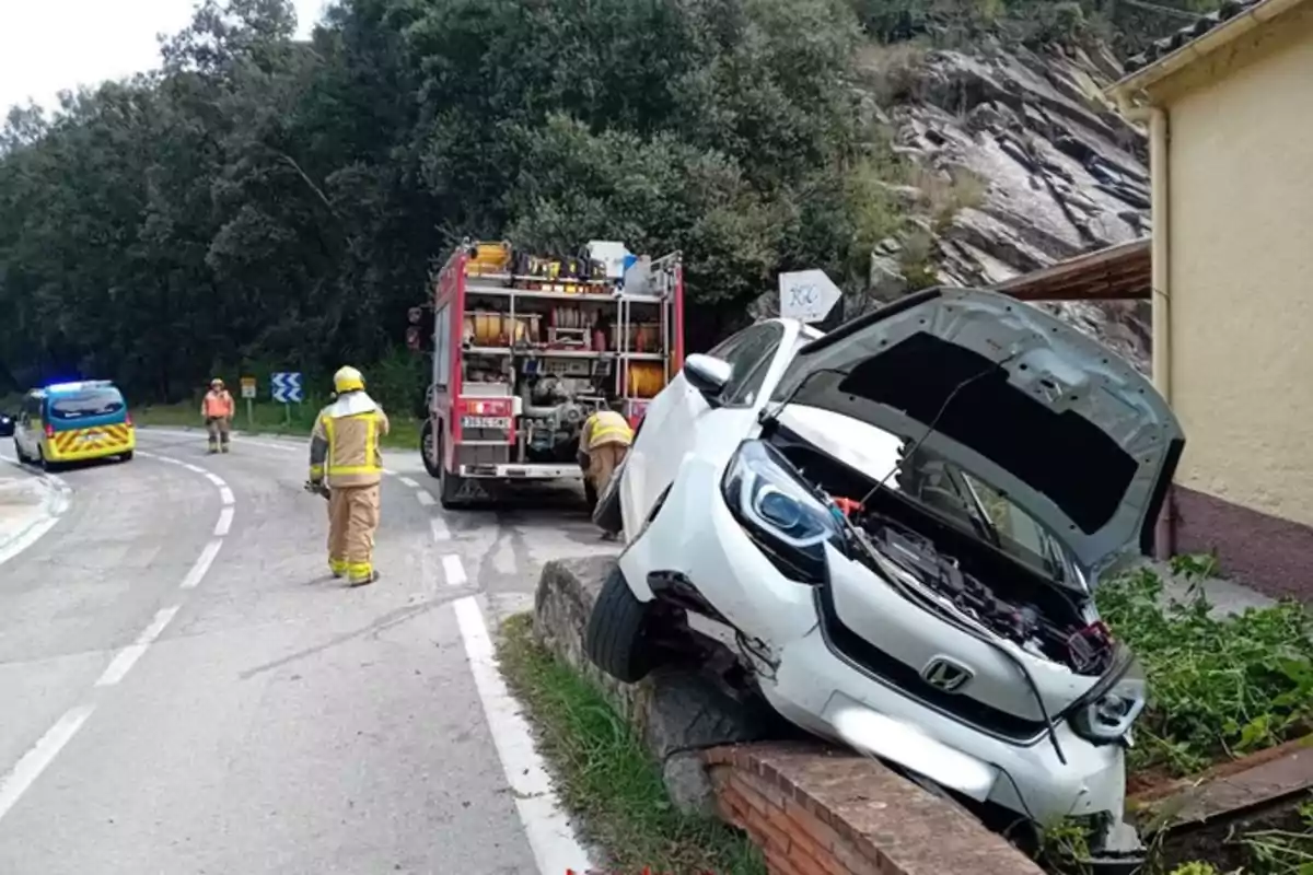 Un coche blanco accidentado sobre un muro al lado de una carretera con un camión de bomberos y personal de emergencia en la escena.