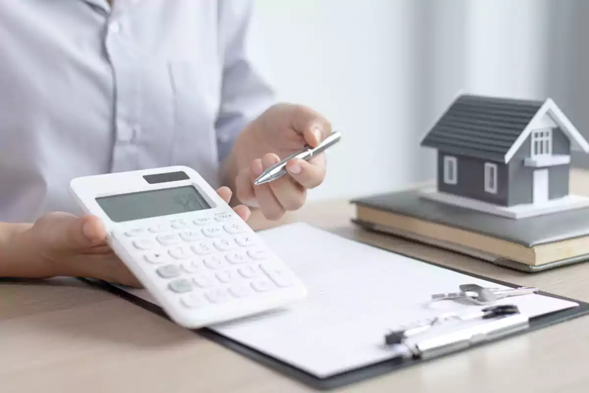 Persona sosteniendo una calculadora y un bolígrafo junto a un modelo de casa sobre un escritorio con documentos.