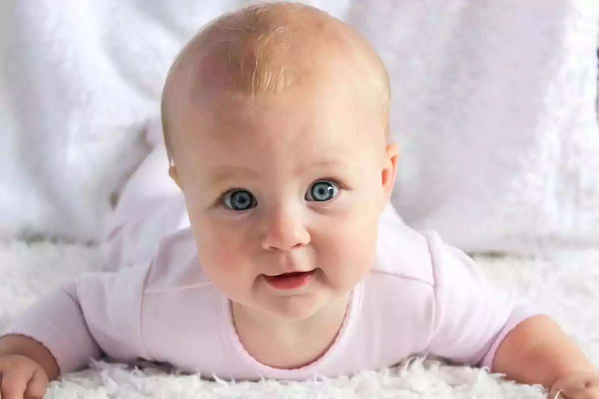 Bebé con ojos azules y cabello rubio acostado boca abajo sobre una manta blanca.