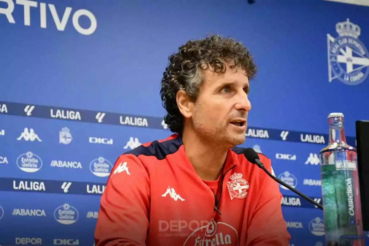 Un hombre con una sudadera roja del Deportivo La Coruña hablando en una conferencia de prensa con un fondo azul que muestra varios logotipos de patrocinadores y de LaLiga.