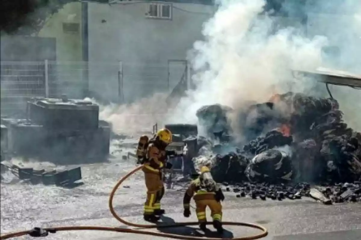 Bomberos combatiendo un incendio con mangueras en una zona industrial con mucho humo.