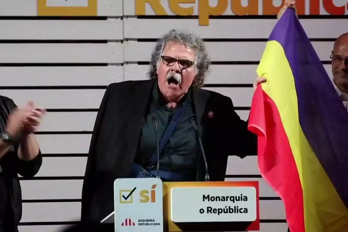 Un hombre con bigote y gafas sostiene una bandera tricolor mientras habla en un podio que tiene un cartel con la opción "Monarquía o República".