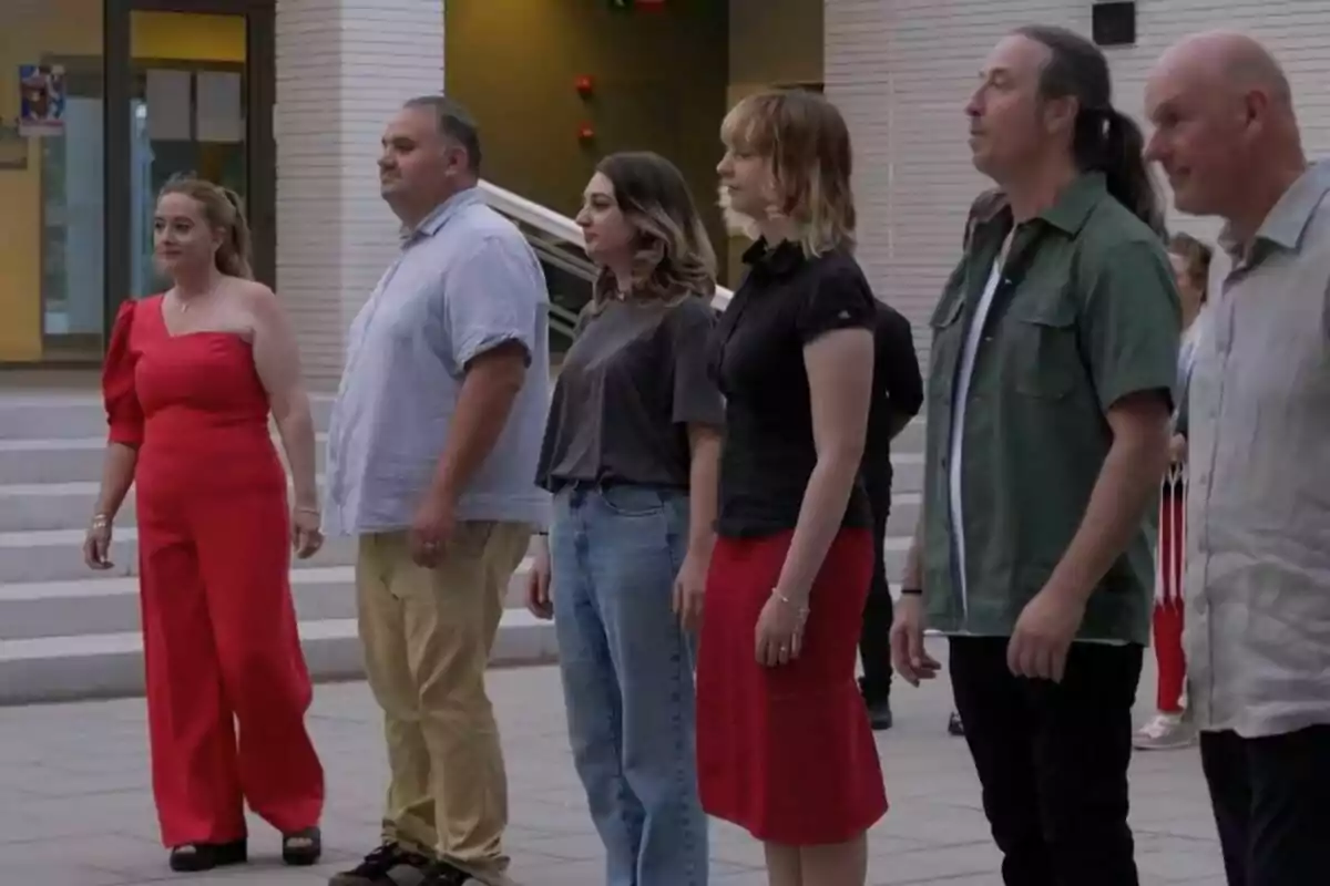 Un grupo de personas está de pie en fila al aire libre, frente a un edificio con escaleras y una entrada iluminada.