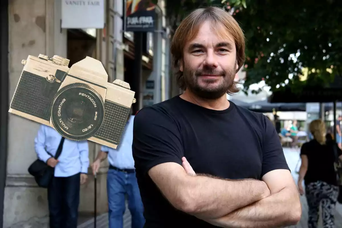 Un hombre con barba y cabello largo, vestido con una camiseta negra, está de pie con los brazos cruzados en una calle concurrida; en la esquina superior izquierda de la imagen hay una cámara fotográfica antigua superpuesta.